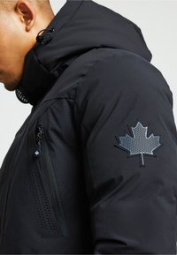 Melnā virsjakā ar iezipjama kabatu, teksturēta auduma un izteiksmīga kļavas lapas logo uz pleca. Iekļauj kapuci un regulējamas auklas.