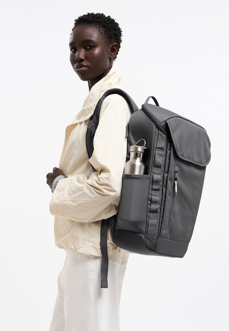 Junge schwarze Frau trägt eine cremefarbene Jacke und weiße Hose und hat einen großen schwarzen Rucksack mit einer Metallwasserflasche in der Seitentasche.