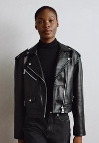 Schwarze Leder-Bikerjacke mit silbernen Reißverschlüssen und Druckknöpfen. Sie hat ein enges, tailliertes Design und klassische Revers. Wird über einem schwarzen Oberteil getragen.