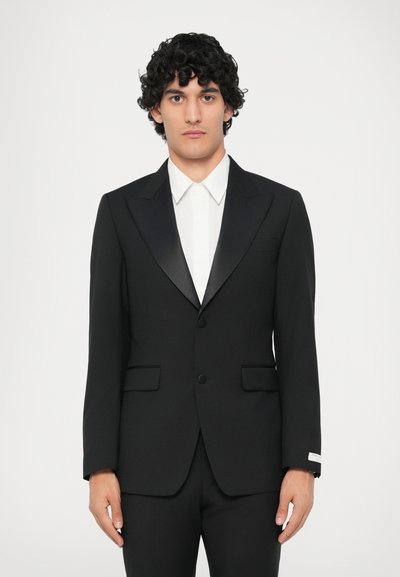 Čierna smokingová bunda so saténovými špicatými klopami, dvoma prednými vreckami, uzáverom na jedno tlačidlo, v kombinácii s bielou košeľou s golierom.