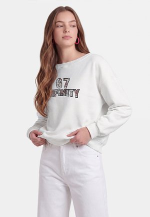 Giovane donna con capelli lunghi indossa una felpa bianca con il testo "67 INFINITY" e jeans bianchi, in piedi contro uno sfondo chiaro e uniforme.