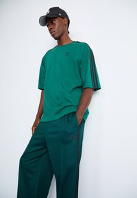 Grønt atletisk outfit bestående af en kortærmet skjorte og matchende bukser, begge med sorte sidelister og et lille logo på brystet.