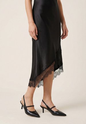 Sort satin midi-nederdel med asymmetrisk blondekant, båret med sorte spidse slingback hæle på en lys baggrund.