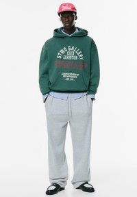 Felpa verde con testo grafico, pantaloni della tuta grigi e scarpe nero slip-on. Include un cappellino rosso e una camicia blu a strati sotto.