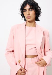 Hellrosa maßgeschneiderte Blazerjacke mit Revers und einem einzigen Holzknopf, kombiniert mit einem passenden, taillierten Oberteil und figurschmeichelnden Hosen.