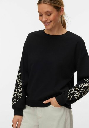 Donna che indossa una maglietta nera a maniche lunghe con motivi floreali bianchi sulle maniche, sorride con gli occhi chiusi, una mano in tasca dei pantaloni chiari.