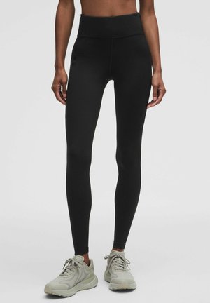 Donna che indossa leggings neri a vita alta e scarpe da ginnastica grigio chiaro, in piedi davanti a uno sfondo chiaro uniforme.