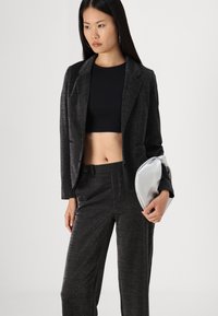 Blazer noir texturé et pantalon assorti, présentant un léger éclat. Souligné par une pochette en argent arrondie, porté avec un top cropped noir.