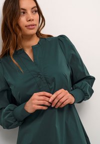Dunkelgrüne Bluse mit hohem Kragen, Knopfverschluss und puffärmel. Glattes, leichtes Material mit lockerer Passform und gerafften Bündchen.