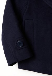 Manteau en laine bleu marine avec des détails de bouton visibles sur les manches. Le tissu semble doux, et les manches présentent une coupe droite avec une poche plaquée.