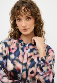 Genomskinlig blus med ett färgglatt blommönster i rosa, blått och svart. Har en ståkrage, knäppningar vid ärmsluten och subtila sömdetaljer.