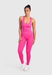 Tajtat pink jumpsuit med tank-aktiga axelband, slät textur och ben i full längd. Bärs med vita träningsskor.