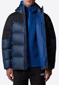 Pufowy czarny i niebieski kurtka z zamkiem błyskawicznym z przodu, elastycznymi mankietami i kapturem. Nosi się ją na niebieskiej teksturowanej koszuli, co tworzy warstwowy wygląd.