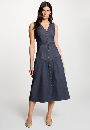 Frau steht und trägt ein ärmelloses, dunkelblaues Jeans-Midikleid mit Knöpfen vorne und beige gewebte flache Schuhe vor einem schlichten Hintergrund.