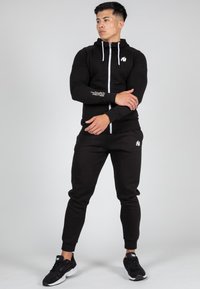 Crna dukserica na zip s bijelim vezicama i logom, u kombinaciji s crnim trenirkama; oboje izrađeno od mekane, teksturirane tkanine.