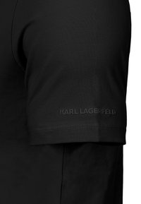 Rukáv černého trička s raženým logem "KARL LAGERFELD" poblíž lemu, zobrazující texturu látky a dvakrát prošitý okraj.