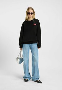 Felpa nera con logo rosa, jeans a gamba larga azzurri chiari e scarpe nere con punta. Tiene una borsa a forma di cuore argentata.