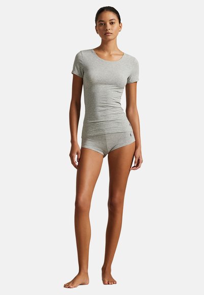 Γυναίκα που στέκεται ξυπόλυτη, φοράει εφαρμοστή ανοιχτογκρι κοντομάνικη μπλούζα και ταιριαστό σορτς μπροστά σε λευκό φόντο.