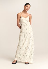 Maksi obleka v krem barvi s črnimi pikami, z tankimi špagetnimi naramnicami, gladko teksturo tkanine in oprijetim krojem.