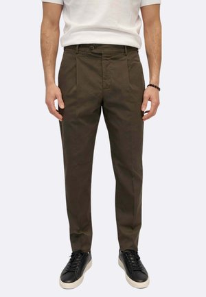 Uomo che indossa pantaloni marroni plissettati, camicia bianca a maniche corte, sneaker nere con suole bianche e un braccialetto nero con perline.