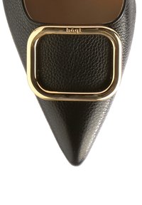 Pumps de piel negra de punta afilada con un destacado acento de hebilla cuadrada dorada, superficie texturizada y un diseño elegante.