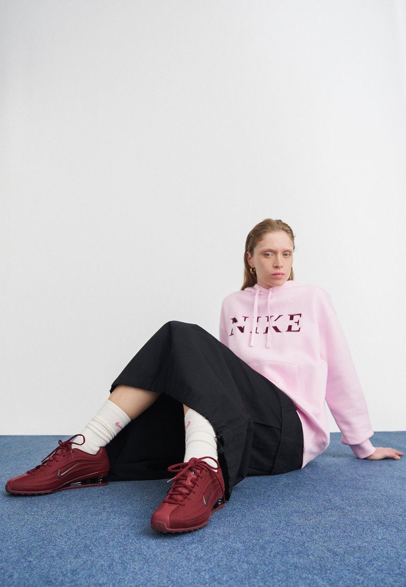 Nike rozā hoody ar logo, apvienota ar melnu svārku. Bordo sporta apavi un krēmkrāsas zeķes ar smalku rakstu. Zils paklājs.