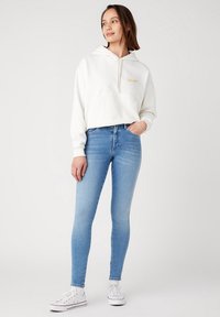 Sudadera blanca con un bolsillo frontal y logotipo bordado, combinada con unos pantalones vaqueros ajustados de color azul claro y zapatillas blancas. Textura suave, ajuste casual.