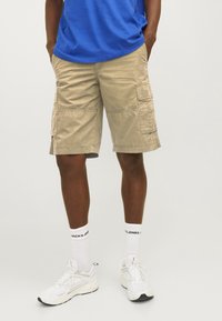 Pantaloni cargo beige con molte tasche, vestibilità rilassata, realizzati in tessuto resistente, abbinati a sneaker bianche e calze corte a coste.