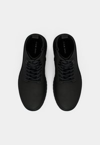 Zapatos negros de tobillo con un acabado suave y mate. Presentan cordones, punta redondeada y una suela de goma robusta. Etiqueta de la marca visible en el interior.