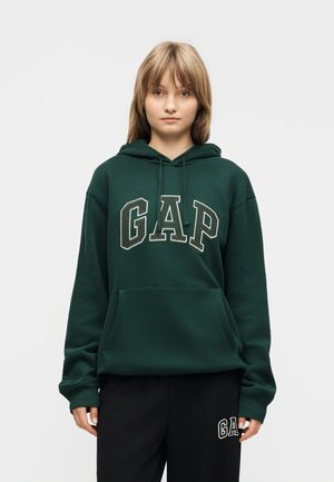 Młoda osoba o jasnobrązowych włosach, ubrana w ciemnozieloną bluzę z kapturem GAP i czarne dresy GAP, stojąca na jasnym, jednolitym tle.