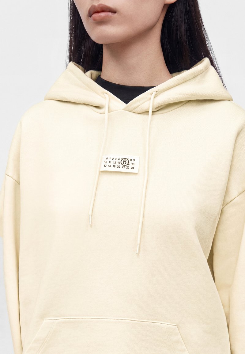 Femme portant un sweat à capuche beige clair avec une petite étiquette rectangulaire comportant des chiffres et un chiffre six encerclé sur la poitrine.