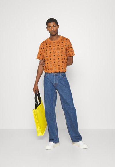 Orange t-shirt med svart logomönster, i kombination med blå jeans. Modell som håller en gul tote bag med svarta handtag. Vita sneakers.