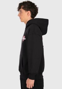 Young man with curly hair wearing a black hoodie with a pink heart graphic on the chest, standing in profile against a plain background.