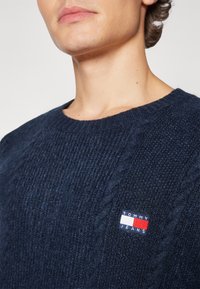 Pull en maille bleu marine en tissu texturé, doté d'un col côtelé et d'un petit badge logo Tommy Jeans sur la poitrine gauche.