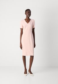 Light pink, fitted dress with short sleeves, V-neckline, and seam detailing. Paired with white, pointed-toe heels. Smooth texture.