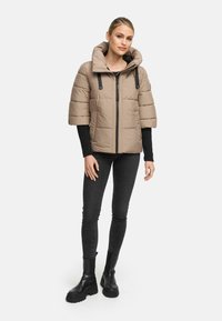 Beige pufferjack met een grote capuchon, korte gewatteerde mouwen en zwarte faux leren accenten, gecombineerd met zwarte skinny jeans en zwarte enkellaarsjes.