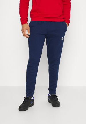 Man die een marineblauwe Adidas trainingsbroek, zwarte sportschoenen en een rode trui draagt, met zijn handen in zijn zakken staand tegen een witte achtergrond.
