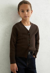 Brun stickad cardigan med V-ringning, prydd med fem svarta knappar, ribbade ärmar och en figursydd midja, tillsammans med mörka denimjeans.