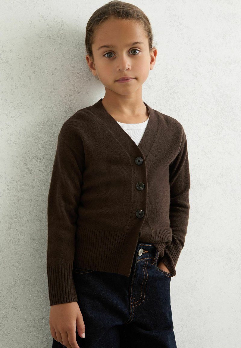 Brun stickad cardigan med V-ringning, prydd med fem svarta knappar, ribbade ärmar och en figursydd midja, tillsammans med mörka denimjeans.