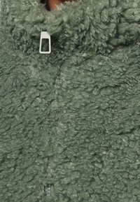 Grön fleecejacka med en texturerad, fluffig yta; har en halv dragkedja, hög krage och ett mjukt, mysigt utseende.