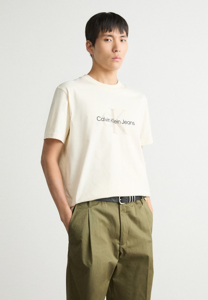 Kreemas kokvilnas T-krekls ar tumši pelēku "Calvin Klein Jeans" logo un stilizētu "CK". Apvienots ar olīvu zaļām plecu biksēm.