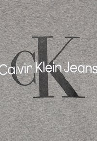 Tessuto grigio con una grande "K" nera e il testo "Calvin Klein Jeans" in bianco, che mostra una texture liscia e un design informale.