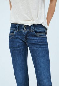 Nära bild av en person som bär mörkblå denimjeans med dubbelknappslåsning och en ljus knuten t-shirt.