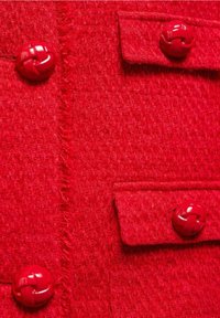 Tissu rouge texturé avec un motif tissé. Comporte quatre boutons rouges au design tressé et deux petites poches. Les bords présentent des détails effilochés.