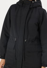 Giacca isolante nera con cappuccio, vita regolabile con cordoncino e due tasche frontali. Texture liscia con polsini elasticizzati e zip frontale.