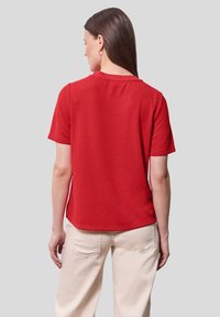 Červené tričko s krátkým rukávem, hladkou texturou a kulatým výstřihem, zkombinované se světlými kalhotami. Jednoduchý a neformální design.