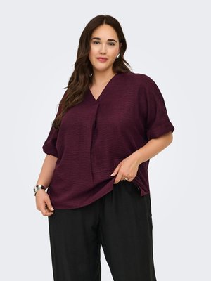 Femme aux longs cheveux bruns portant un t-shirt ample violet foncé à col en V et un pantalon noir, debout devant un fond clair uni.