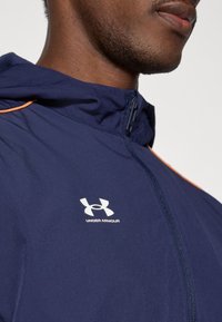 Μπλε ναυτικού χρώματος ζακέτα με φερμουάρ και πορτοκαλί λεπτομέρειες, διαθέτει κουκούλα και λευκό λογότυπο Under Armour στο στήθος, κατασκευασμένη από ελαφρύ υλικό.