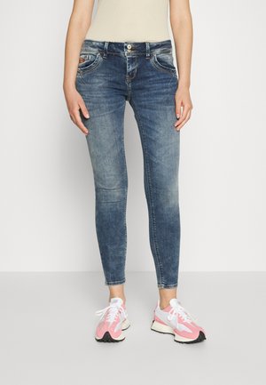 Kvinde iført falmede blå skinny jeans og hvide og lyserøde løbesko, stående mod en ensfarvet hvid baggrund.