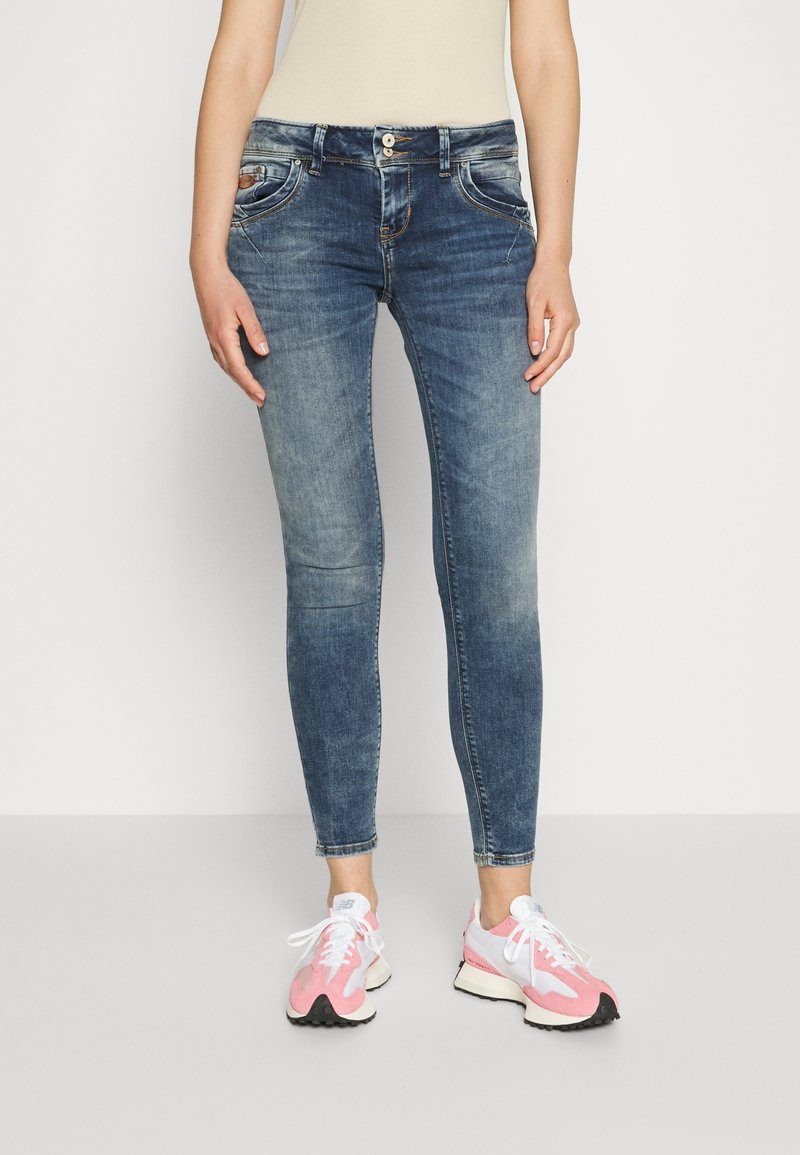 Femme portant un jean skinny bleu délavé et des chaussures de course blanches et roses, debout sur un fond blanc uni.